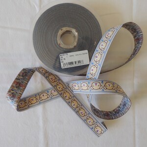 May include: A roll of white ribbon with a blue and brown pattern featuring cartoon bear faces. The ribbon is wrapped around a spool with a label that reads "Art 0179 - 25 mm - 100% Polyester - 02".