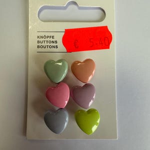 Peut inclure: Un paquet de six boutons en forme de cœur de différentes couleurs pastel : vert, pêche, rose, violet, gris et vert citron. Le carton indique "KNÖPFE BUTTONS BOUTONS".