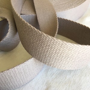 Può includere: Un'immagine ravvicinata di un rotolo di fettuccia di cotone beige, un tipo di nastro in tessuto resistente. La fettuccia è tessuta con un motivo testurizzato ed è piegata per mostrare la larghezza e lo spessore del materiale.