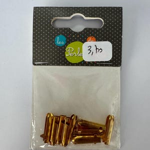 Op de afbeelding: Een pakket goudkleurige metalen fournituren, waarschijnlijk voor sieraden of knutselen. De fournituren zitten in een doorzichtige plastic zak met een zwart-witte stippen header. De header heeft de tekst "les Perle de" en een prijskaartje.