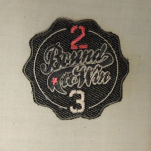 Pode incluir: Etiqueta bordada preta e branca com o texto "Bound to Win" em uma fonte estilizada. Os números "2" e "3" estão em vermelho e branco, respectivamente.