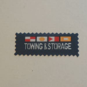 Könnte beinhalten: Ein blaues und weißes rechteckiges Schild mit einem gewellten Rand. Das Schild hat vier kleine Flaggen in den Farben Rot, Weiß und Gelb. Der Text auf dem Schild lautet "Towing & Storage".