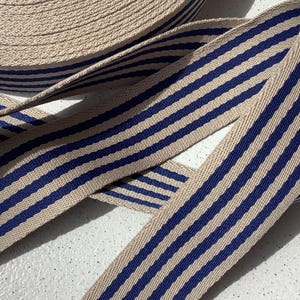 Puede incluir: Un rollo de cinta a rayas beige y azul marino. La cinta está enrollada firmemente, con una parte desenrollada, mostrando las rayas azul marino sobre el fondo beige. La cinta parece estar hecha de tela tejida.