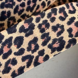 May include: Close-up of a fabric with a leopard print pattern. The fabric has a tan background with dark navy blue and pink spots. The fabric is folded over, showing the pattern in detail.
