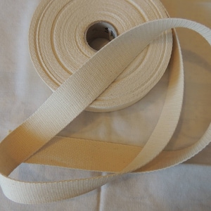 May include: A roll of natural cotton webbing, a type of strong, woven fabric used for straps, belts, and other applications. The webbing is rolled up and a length of it is laid out on a white surface.