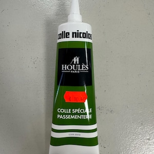 Peut inclure: Un tube de colle Colles Nicolas avec un embout blanc et une étiquette verte et noire. L'étiquette porte le texte "HOULES PARIS" et "COLLE SPÉCIALE PASSEMENTERIE". Le tube présente un motif à rayures blanches et vertes.