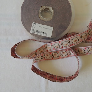 Peut inclure: Un rouleau de ruban beige avec une ligne rouge et un motif répété de visages d'ours bruns. Le ruban mesure 15 mm de large et est composé de 100 % de polyester. L'étiquette sur la bobine indique "Art 9175, 15 mm, 100 % Polyester".
