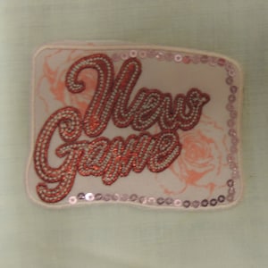 Könnte beinhalten: Ein rosa-weißer Stoff-Patch mit rosa Pailletten am Rand. Der Patch hat einen roten gestickten Text mit der Aufschrift "New Game".