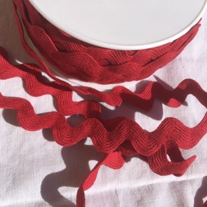 Puede incluir: Cinta de adorno roja en zigzag en un carrete blanco. El adorno es un patrón ondulado en zigzag.