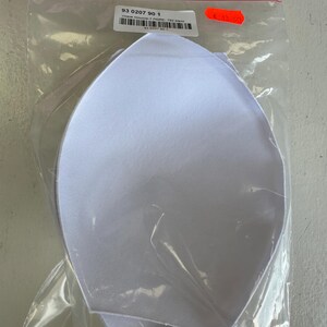 Puede incluir: Un par de copas de sujetador blancas en una bolsa de plástico transparente. Las copas son de un material suave y acolchado con forma curvada. Una etiqueta con el número "93 0207 90 1" es visible.