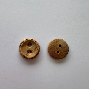 Op de afbeelding: Twee ronde houten knopen. Eén knoop heeft een decoratieve gestikte rand en twee gaten. De andere knoop is effen met twee gaten. Beide knopen zijn lichtbruin met zichtbare houtnerf.