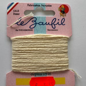 Puede incluir: Un carrete de hilo color crema etiquetado "le Zaufil" con el texto "Fabrication française" y "depuis 1946" en una tarjeta rosa y blanca. El hilo está bien enrollado alrededor del carrete.