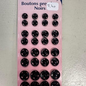Puede incluir: Una tarjeta rosa con un surtido de 32 botones de presión negros de varios tamaños, de 9 mm a 15 mm. La tarjeta tiene texto en francés, incluyendo "Boutons Noirs" y "Couture Loisirs."