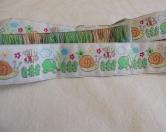 May include: Light blue ribbon with embroidered garden scene featuring snails, bees, frogs, and flowers. The ribbon has a green fringe along the bottom edge. The design includes a green picket fence and a blue sky.