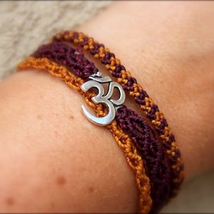 May include: A close-up of a wrist wearing three braided bracelets. The bracelets are made of brown and burgundy cord and feature a silver Om charm.