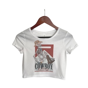 Puede incluir: Top corto blanco con un gráfico de un vaquero esqueleto con sombrero y botas, con el texto "COWBOY killers" en un diseño rojo y negro.