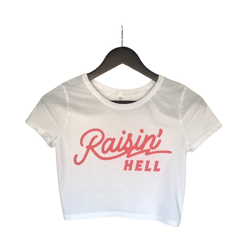 Puede incluir: Camiseta blanca recortada de manga corta con un gr&aacute;fico rosa que dice "Raisin' Hell"