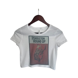 May include: White cropped t-shirt with a graphic print of a vintage poster for the Pendleton Round-Up. The poster features a cowboy riding a bucking bronco. The text on the poster reads "Pendleton Round-Up Pendleton, Oregon".