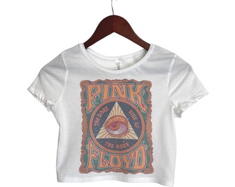 May include: White cropped t-shirt with a vintage-style graphic of Pink Floyd's "The Dark Side of the Moon" album cover. The graphic features a triangle with an eye inside, surrounded by text and a decorative frame.