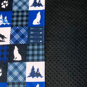 Peut inclure: Une couverture patchwork bleue et noire avec des motifs de loups blancs, d'empreintes de pattes et de scènes de forêt. Le verso est noir et texturé.