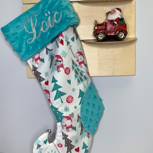 May include: A Christmas stocking with a teal cuff embroidered with the name "Loic" in silver thread. The stocking body features a pattern of deer, trees, and snowflakes on a white background, with a teal accent. A small silver bell hangs from the toe.