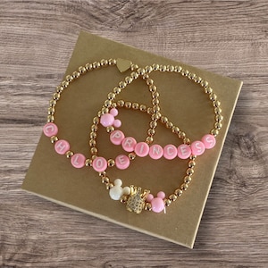 May include: Three gold beaded bracelets with pink beads spelling out the names "Chloe", "Princess", and a crown charm.
