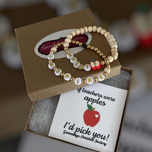 May include: Two bracelets with wooden beads and gold beads spelling out "INSPIRE" and "MRS" with a red apple charm. The bracelets are in a brown box with a white card that says "If teachers were apples I'd pick you!" and "Gwendolyn Christine Jewelry".