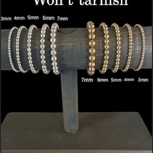 May include: A wooden display stand with six gold and silver bracelets. The bracelets are made of small beads and are labeled with their size in millimeters: 3mm, 4mm, 5mm, 6mm, and 7mm. The text "Won't tarnish" is displayed at the top of the image.