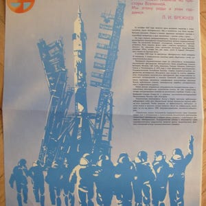 May include: A vintage poster with a blue and white illustration of a rocket on a launch pad, with a group of figures in the foreground. The poster has text in Russian.