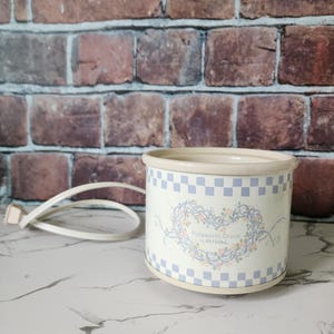 May include: A white and blue Potpourri Crock by Rival. The crock has a floral heart design and a blue checkered pattern around the top. It has a cream-colored rim and base. A white power cord is attached.