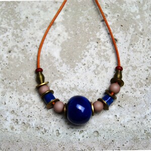 Könnte beinhalten: Eine handgefertigte Halskette mit einer großen, tiefblauen Mittelperle. Die Halskette hat eine leuchtend orangefarbene Kordel, die mit Messing-, rosa- und blauen Perlen akzentuiert ist. Das Design ist vor einem strukturierten, hellgrauen Hintergrund platziert.