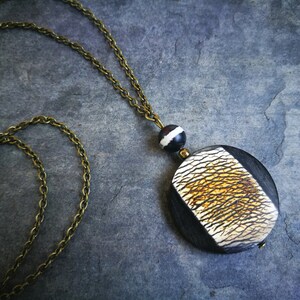 May include: A long necklace with a brass chain and a large, round pendant. The pendant features a black wooden base with a textured, cream and gold-colored inlay. A small black and white bead sits above the pendant.