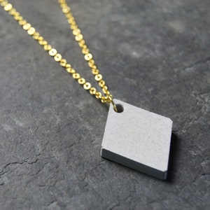 Könnte beinhalten: Halskette mit einem geometrischen Anhänger in Form eines hellgrauen Diamanten, der an einer zarten goldfarbenen Kette hängt. Der Anhänger hat oben ein kleines Loch. Die Halskette wird auf einer strukturierten grauen Oberfläche präsentiert.