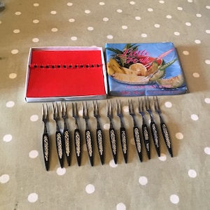 Puede incluir: Un juego de tenedores de fruta vintage con mangos negros y detalles florales dorados, dispuestos sobre una mesa. Los tenedores están junto a una caja roja y una caja con las palabras "Little Forks" y una ilustración de fruta.