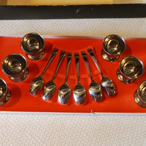 Puede incluir: Un juego de ocho hueveras y cucharas plateadas dispuestas en una caja rectangular con interior naranja. Las cucharas son curvas y las hueveras tienen base de pedestal. La caja tiene borde negro y exterior blanco.