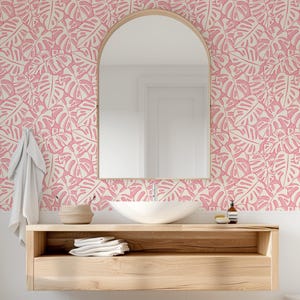 Op de afbeelding: Een badkamer met een lichte houten wastafel, een witte waskom en een grote boogvormige spiegel. De muur is bedekt met roze en crèmekleurig behang met tropische bladeren. Een witte handdoek hangt ernaast.