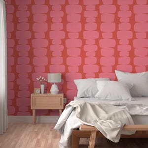 Op de afbeelding: Een slaapkamer met een wit bed en witte beddengoed. De muur achter het bed is bedekt met een rood en roze geometrisch patroon behang. Een houten nachtkastje met een lamp staat naast het bed.