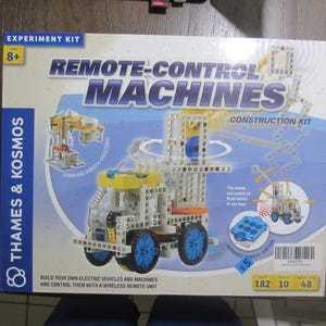 May include: A Thames & Kosmos Remote-Control Machines construction kit box featuring a white, yellow, and blue buildable vehicle with wheels and gears. The packaging displays text including "Experiment Kit," "Ages 8+," and "Build your own electric vehicles."
