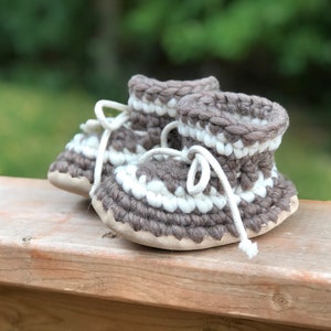 May include: A pair of brown and white knitted baby booties with white laces. The booties are sitting on a wooden surface.