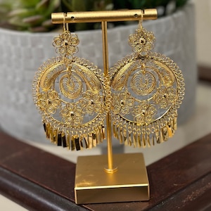 May include: Gold filigree earrings with a floral and fringe design. The earrings are displayed on a gold stand, showcasing their intricate details. The earrings have a circular shape with delicate floral accents and dangling fringe.