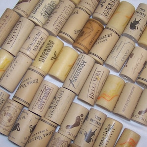 May include: A collection of cork wine bottle stoppers with various winery labels and names, including Bella Sera, Edna Valley, Georges Duboeuf, and Meridian. The corks are made of natural cork and have a variety of colors and textures.