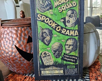 Op de afbeelding: Een zwart houten bord met een groene en witte poster die adverteert voor een monsterfilm genaamd "The Monster Squad: Spook-O-Rama". De poster toont een monster met tanden, een weerwolf en een mummie. Het bord bevat ook de tekst "Creature Features Presents" en "Cast & Crew Reunion!!".