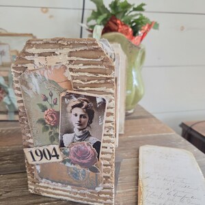 May include: A vintage-style tag with a textured, corrugated cardboard base. It features a sepia-toned portrait of a woman, a rose illustration, and the number "1904". The tag is embellished with layered paper elements.