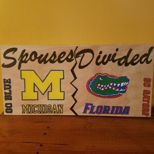 May include: A wooden sign with the text "Spouses Divided" painted in black. The sign is split in half, with a yellow "M" for Michigan on the left and a green alligator for Florida on the right. The text "Go Blue" is on the left and "Go Gators" is on the right.