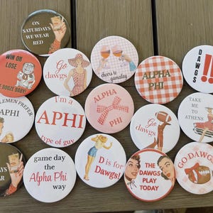 Puede incluir: Una colección de botones redondos con varios diseños y texto. Algunos botones presentan frases como "On Saturdays We Wear Red" y "Go Dawgs". Otros botones muestran imágenes de mujeres, balones de fútbol y el texto "Alpha Phi".
