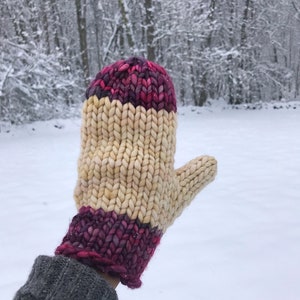 May include: A knitted mitten with a cream coloured body and a pink and purple cuff. The mitten is held up against a snowy background.