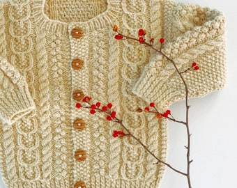 May include: Cream-colored, hand-knitted cardigan with a cable-knit design and wooden buttons. The sweater features intricate patterns and a round neckline. A sprig of red berries adds a pop of color. This cozy, textured garment is perfect for cooler weather.