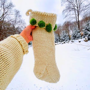 Puede incluir: Calcetín navideño tejido de color crema con pompones verdes. El calcetín se sostiene sobre un fondo nevado con árboles. El puño del calcetín está decorado con pompones verdes.