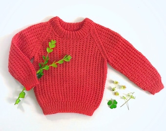 May include: A coral-colored knitted sweater with a ribbed texture. The sweater has a crew neck and long sleeves.
