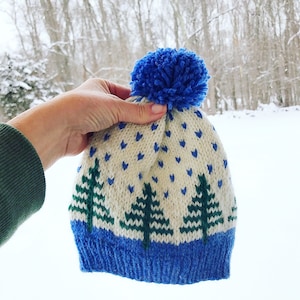 May include: A white knitted hat with a blue pom pom and green pine tree designs. The hat has a blue brim and a white background with blue hearts.